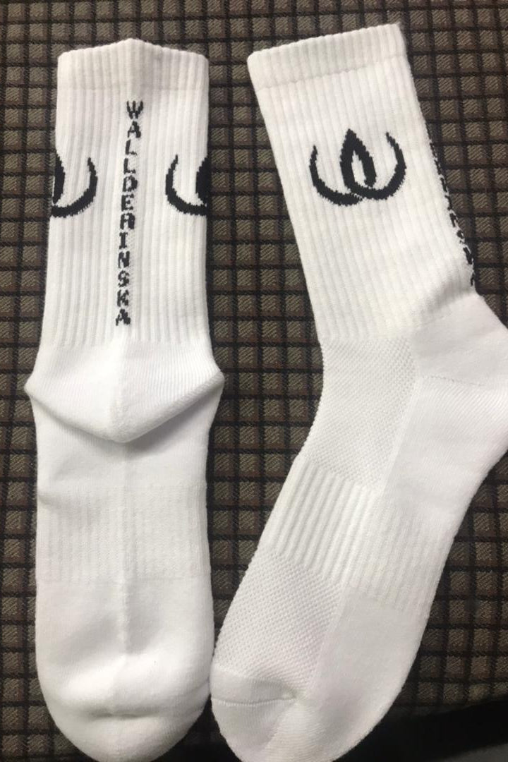 Une paire de chaussettes blanches avec logo noir et écusson le long de la tige, sur fond sombre.