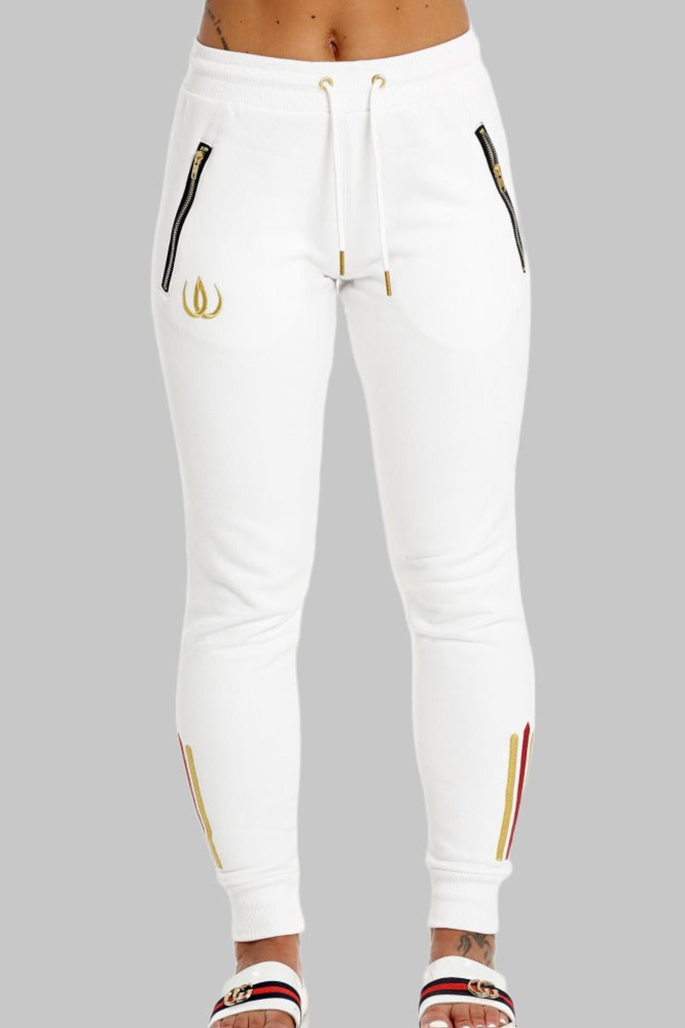 Deux poches zippées dorées sur le jogging blanc, cordons à embouts dorés et logo doré sur la jambe.