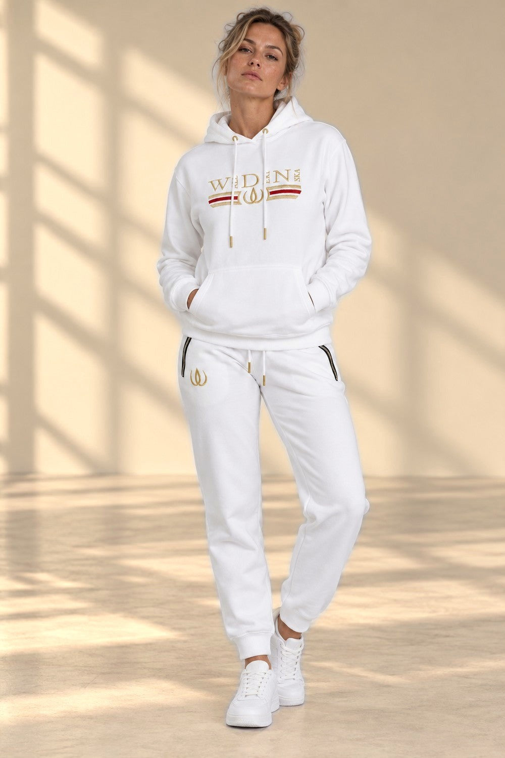 Le modèle dans l'ensemble Deluxe blanc, hoodie et joggers avec fermetures aux poches et logo doré.
