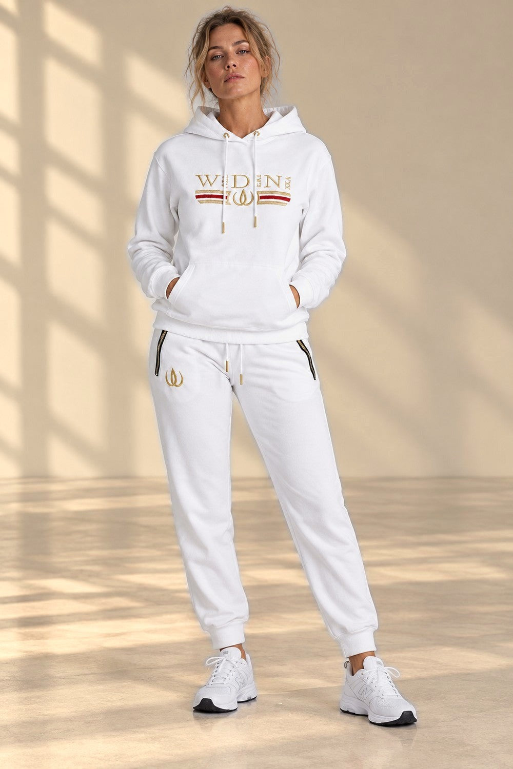 Deluxe Hoodie Blanc et Deluxe Joggers Blanc en pied, avec le logo doré sur la cuisse et des sneakers blancs.