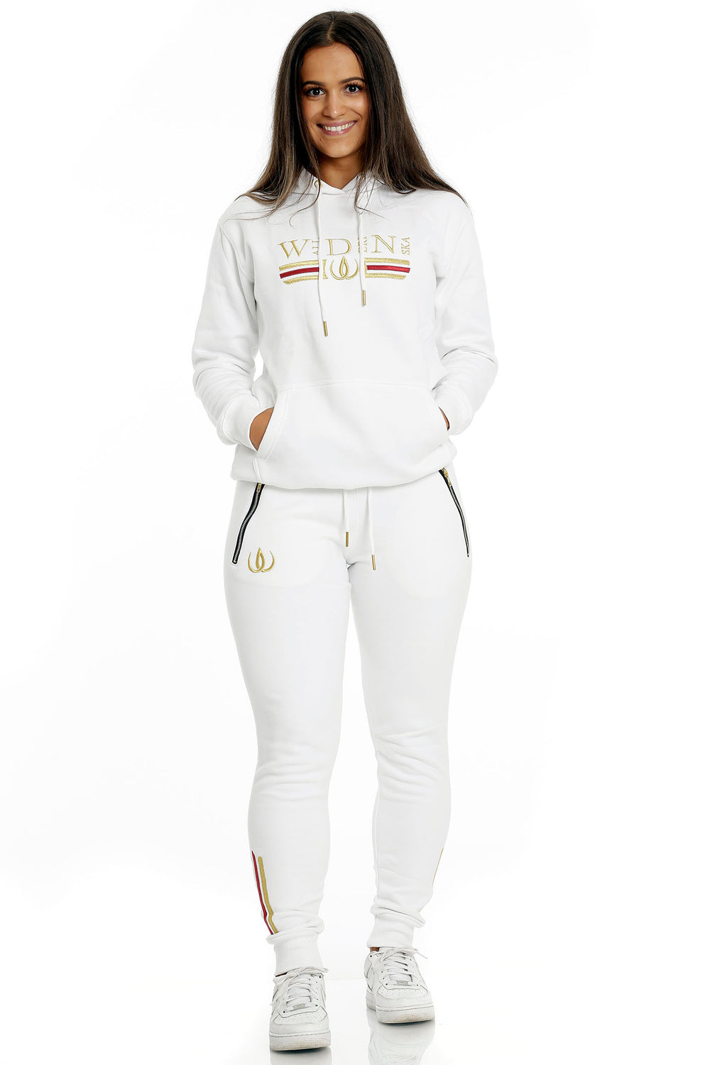 Debout en jogging blanc à logo doré et hoodie blanc à broderie dorée, porté avec des baskets blanches.