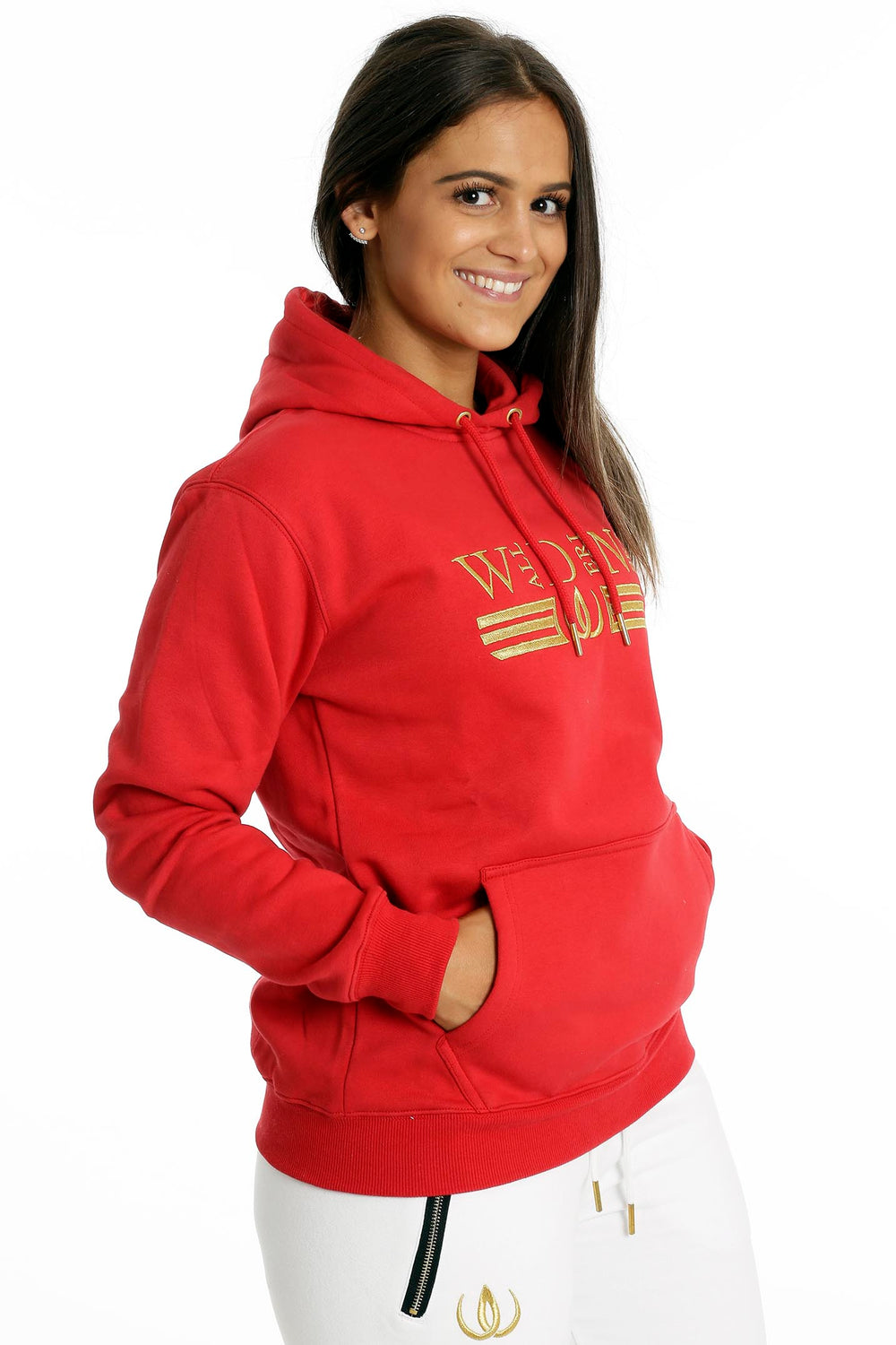 Les mains reposent dans la poche kangourou du hoodie rouge. En dessous, un jogging blanc à zip doré.
