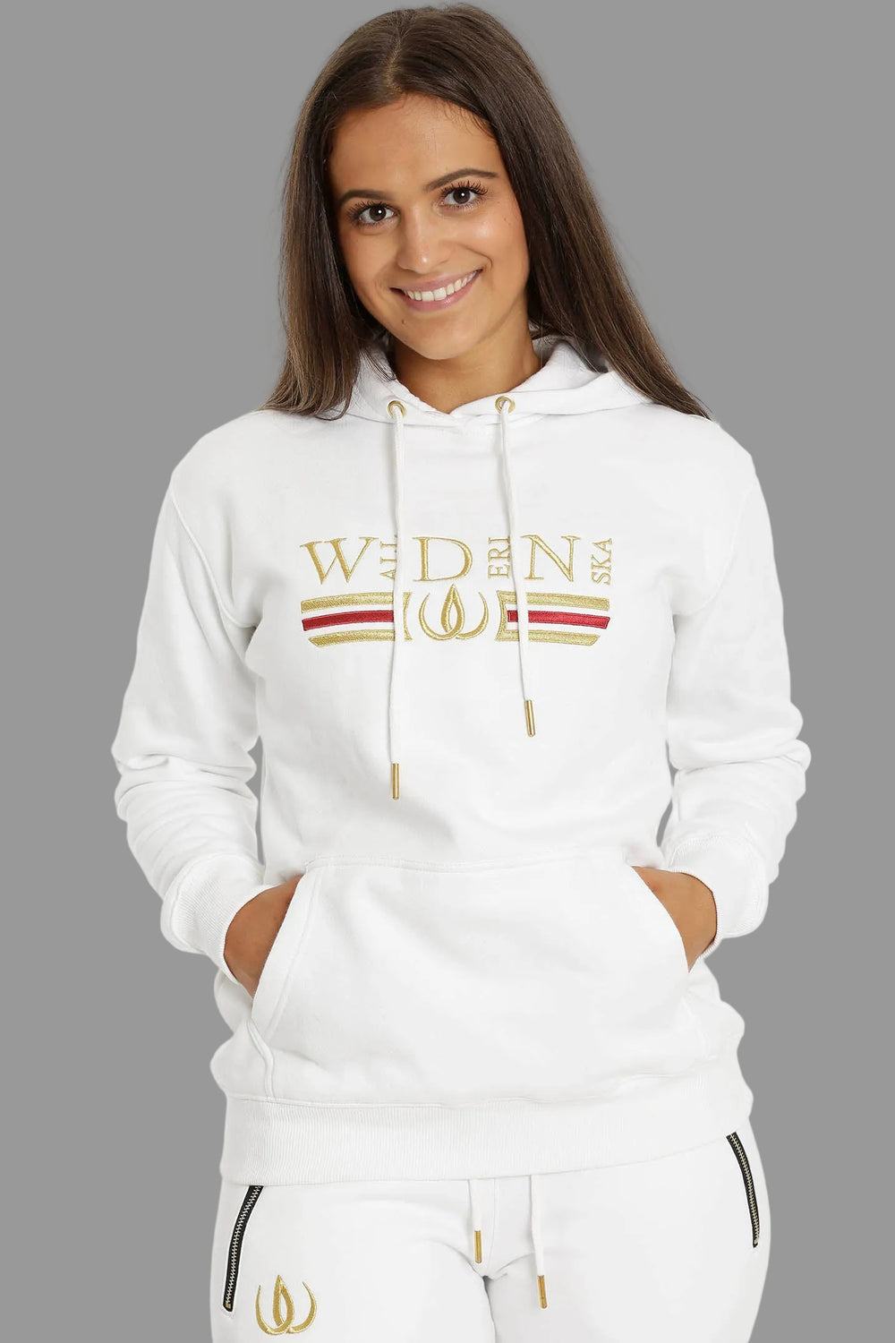 Sweat à capuche blanc avec broderie dorée et rouge sur la poitrine, porté avec des joggers blancs à logo doré.
