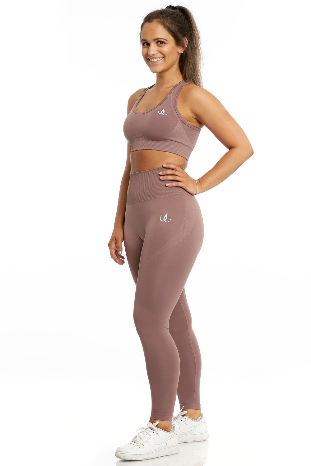 Brassière de sport et legging dans le même ton brun rosé. Le mannequin pose une main sur la hanche, en chaussures blanches.