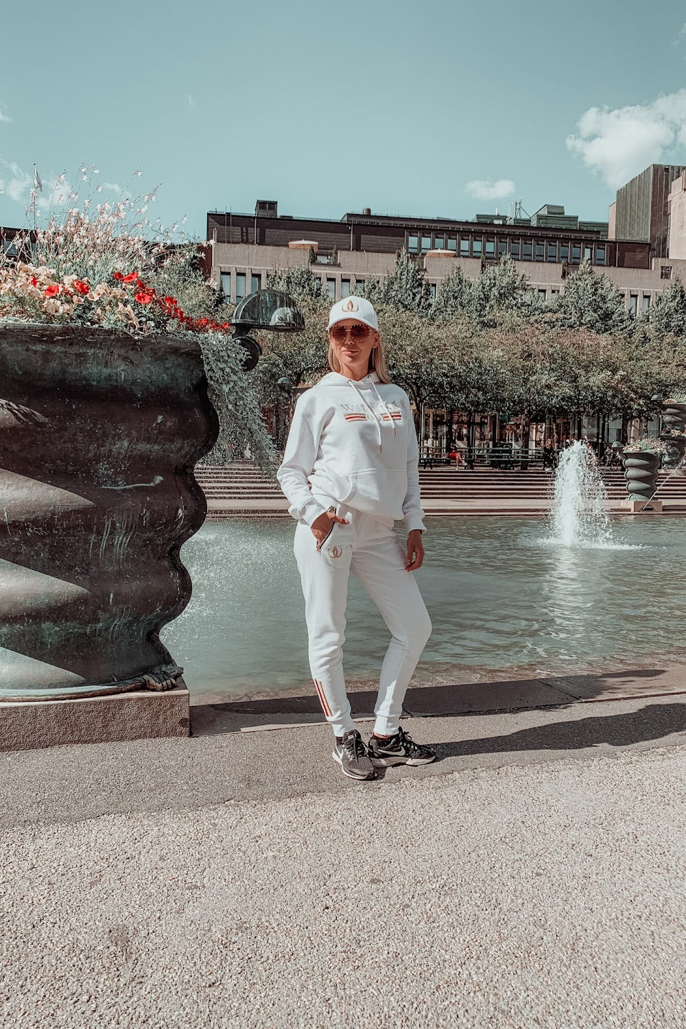 Devant la fontaine, le modèle se tient en hoodie blanc, jogging blanc et casquette blanche.