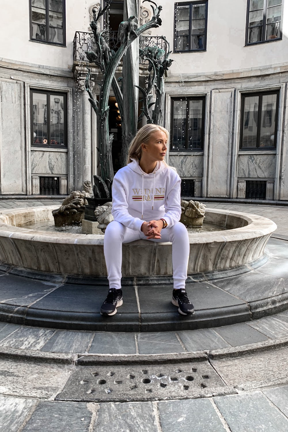 Sur le rebord de la fontaine, le modèle est assis en hoodie blanc, avec une statue en arrière-plan.
