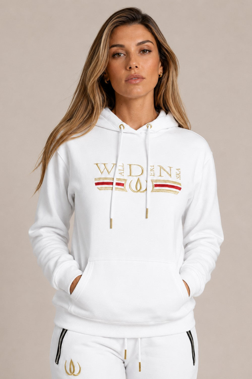 Hoodie Deluxe blanc avec broderie dorée et bande rouge sur la poitrine, porté avec un jogging Deluxe blanc au logo doré.
