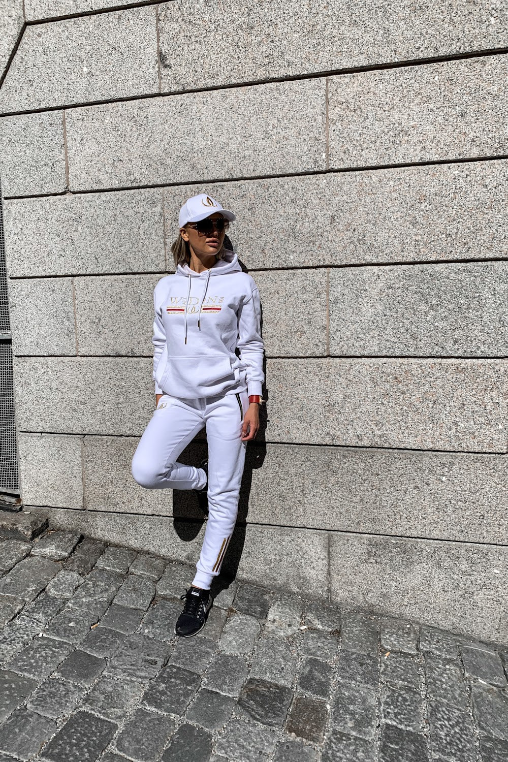 Contre le mur de pierre, le modèle s'appuie avec un pied relevé, en hoodie blanc et jogging blanc.