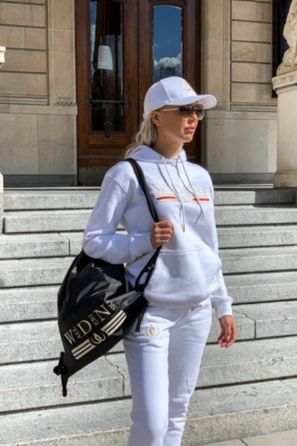Sur les marches devant un bâtiment, le hoodie blanc est porté avec une casquette et le Deluxe Sac à Dos Noir sur l'épaule.