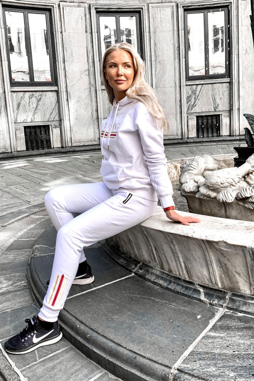 Le mannequin est assis près d'une fontaine en ville, en hoodie blanc et jogging blanc à rayure dorée.