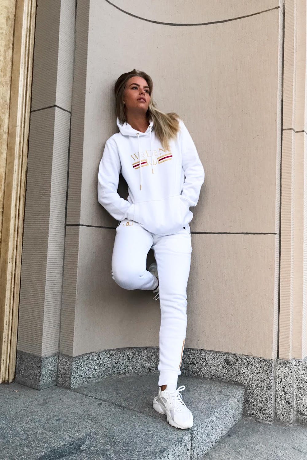 Contre le mur de la maison, le modèle se tient en hoodie blanc et jogging blanc, avec des baskets blanches.