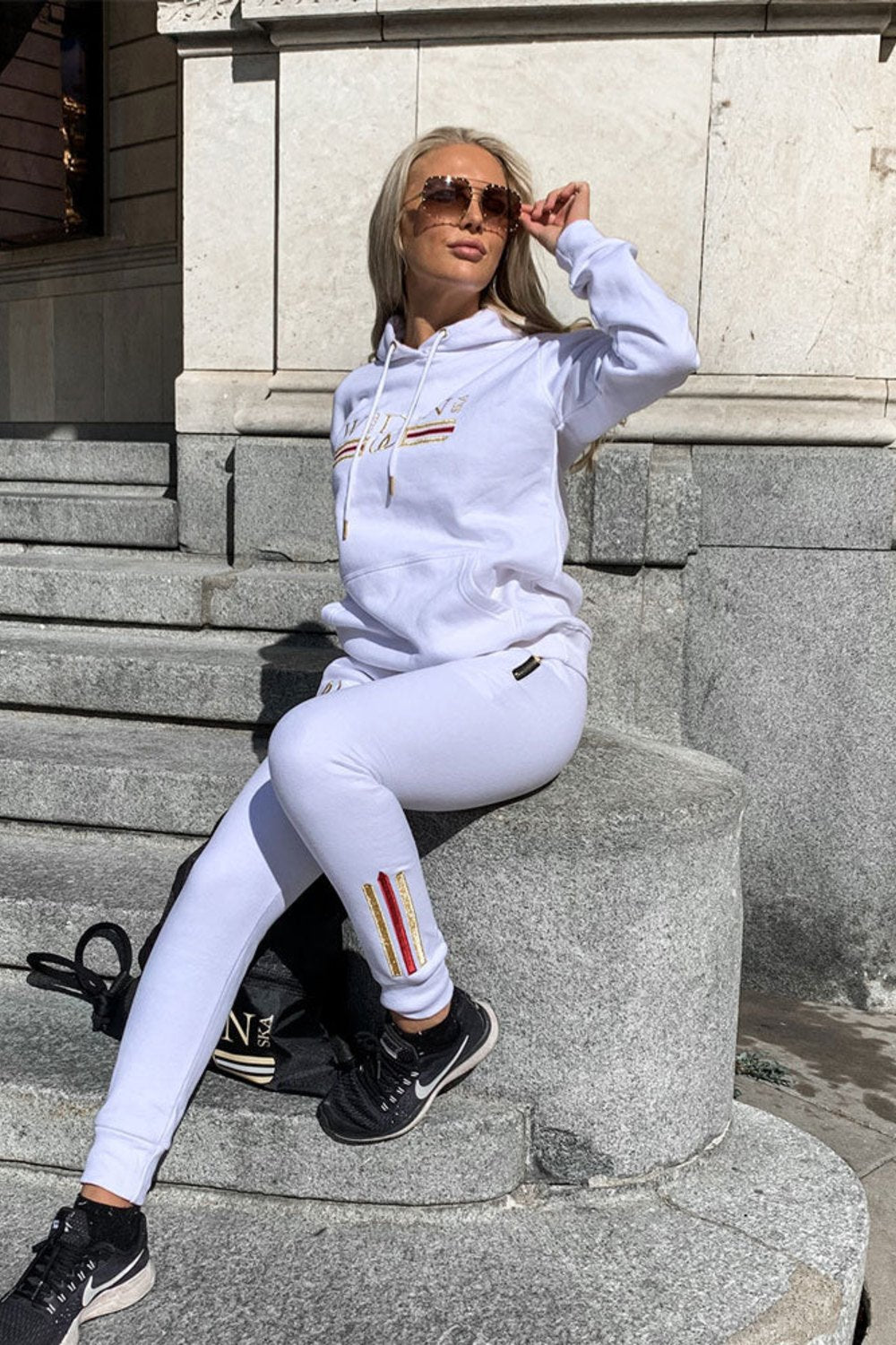 Sur un escalier de pierre en ville, le mannequin porte un jogging blanc à rayure dorée à la cheville et un hoodie blanc.