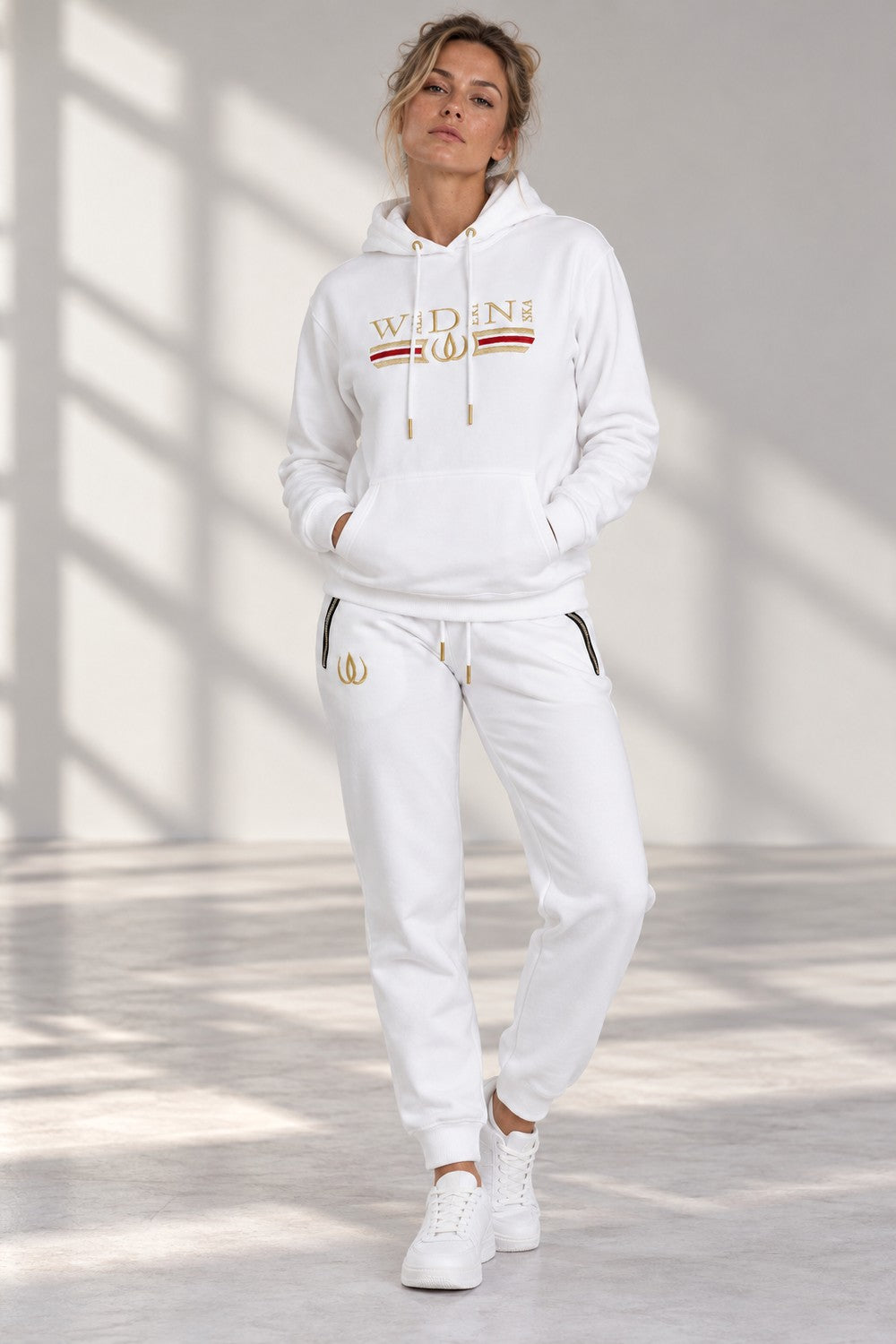 Le modèle dans l'ensemble Deluxe blanc, hoodie et joggers avec fermetures aux poches et logo doré.
