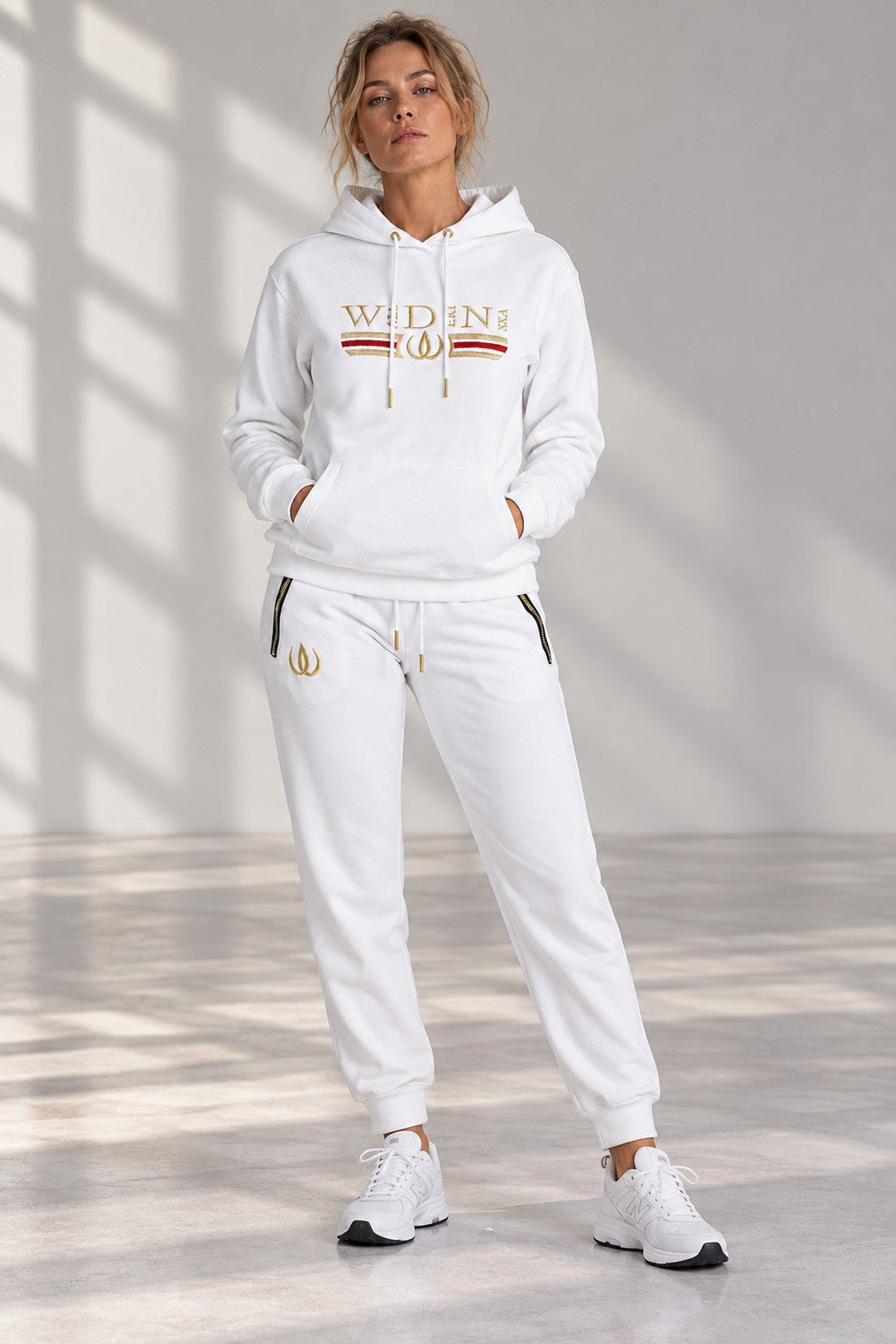 Deluxe Hoodie Blanc et Deluxe Joggers Blanc en pied, avec le logo doré sur la cuisse et des sneakers blancs.