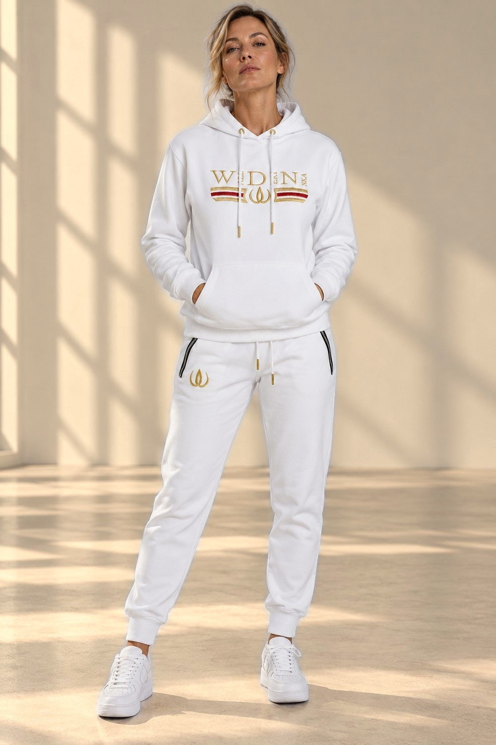 Deluxe Joggers Blanc avec logo doré sur la cuisse et fermetures aux poches, porté avec le hoodie assorti.