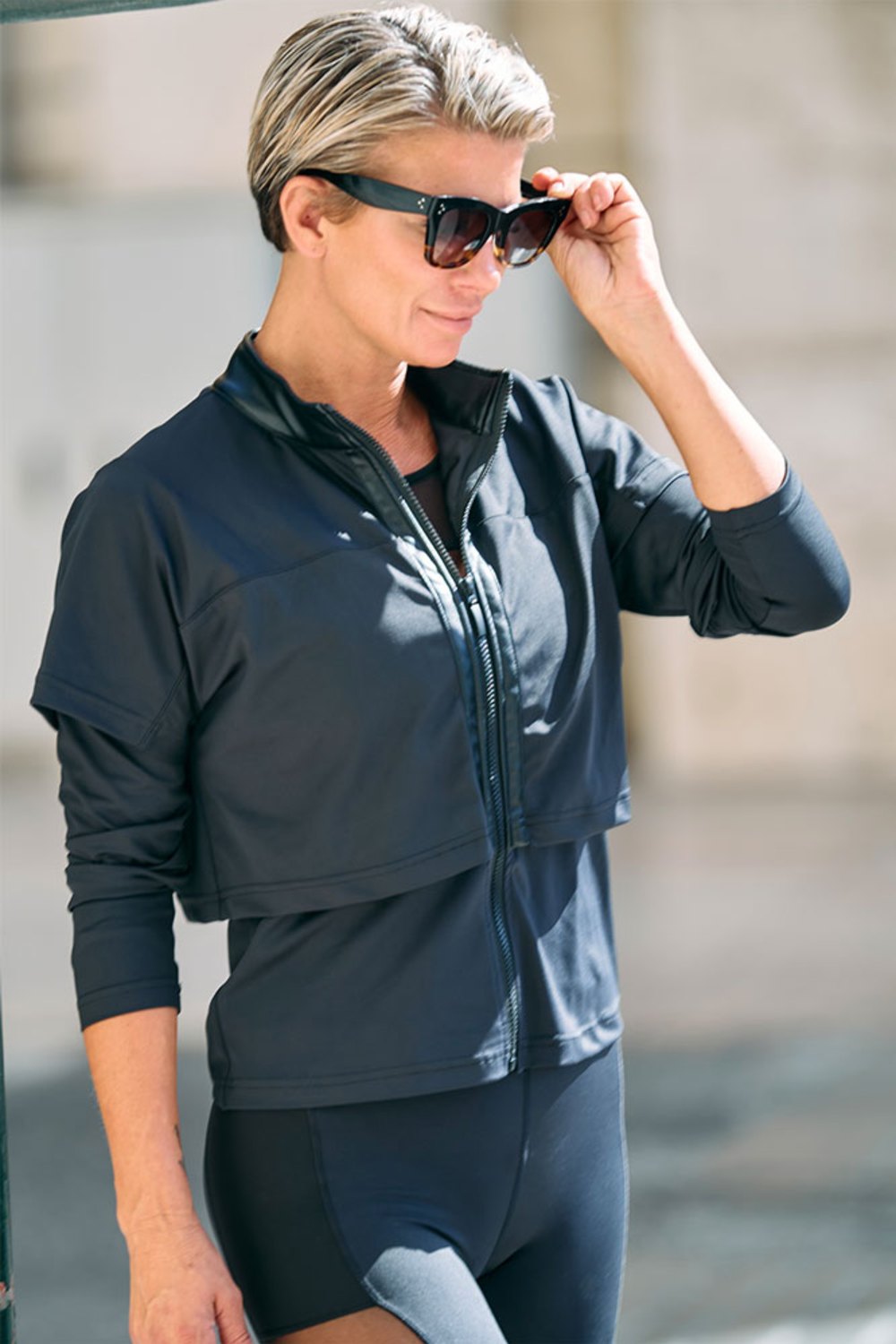 Zip à moitié ouvert sur la veste noire, tandis que le mannequin soulève ses lunettes de soleil.