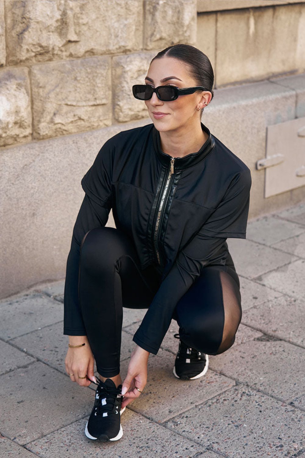 Au bord du trottoir, le mannequin accroupi lace sa chaussure, dans la veste noire et un legging noir.