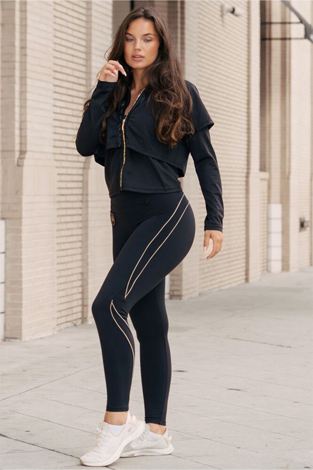 Devant le mur de briques, le mannequin se tient dans la veste et un legging noir aux lignes dorées.