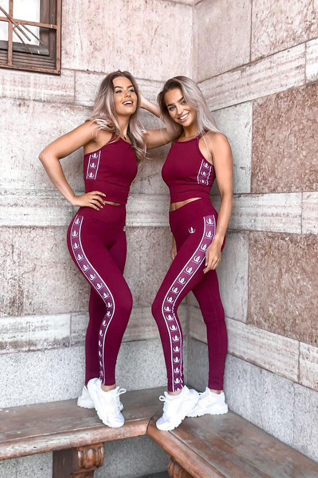 Deux modèles appuyés contre un mur en pierre en crop tops bordeaux et leggings à bandes logo blanches.
