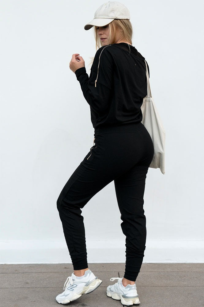 Le dos du pull avec la fermeture éclair dorée sur l'épaule. Le mannequin porte une casquette, un pantalon et un tote bag.