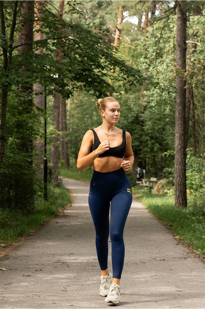 Course sur un chemin forestier en legging bleu marine à rayures dorées et brassière de sport noire.