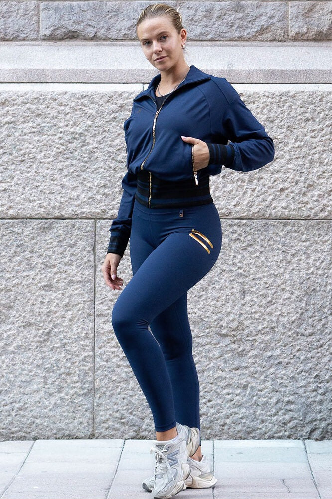 Devant le mur de granit, le legging bleu marine apparaît avec une veste ouverte à fermetures dorées.